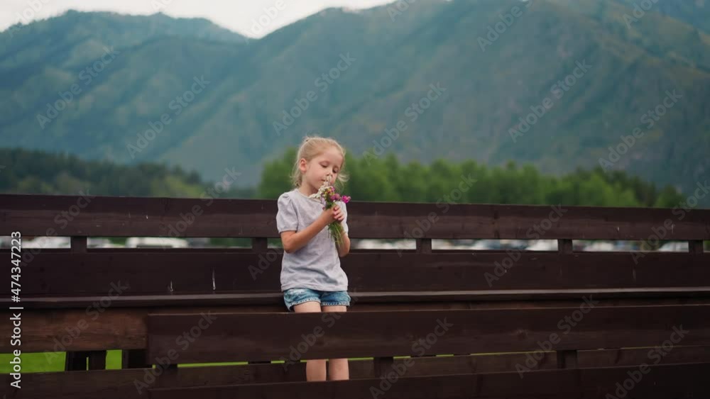 Cute little girl smells small bouquet of windflowers leaning on wooden bench against foggy mountain silhouettes on spring day slow motion