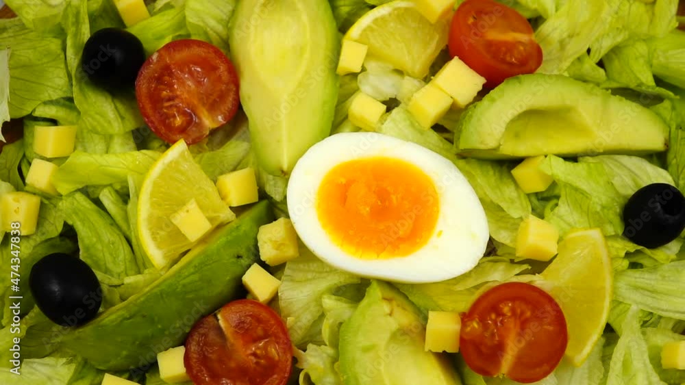 Avocado and lemon slices, half a boiled soft-boiled egg, black olives, slices of cheese and cherry tomatoes lie on iceberg lettuce leaves, slow motion. Festive, vitamin green salad is on the platter
