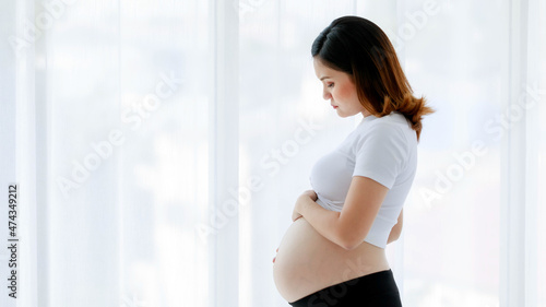 Free Download Asian young stressed depressed upset sick unhappy female pregnancy mother standing alone thinking worrying about unborn baby holding touching naked belly tummy stomach in front curtain background - GFXLoad