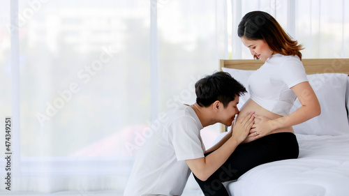 Asian young happy loving supporting comforting soothing husband sitting smiling kissing lovely unborn baby in beautiful pregnancy wife naked big belly tummy stomach sitting on bed in bedroom at home