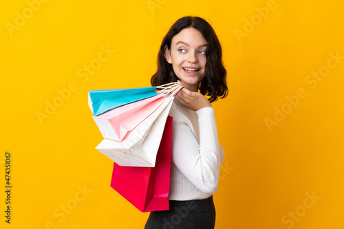Teenager Ukrainian girl isolated on yellow background holding shopping bags and looking back