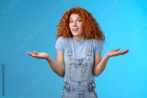 Who knows. Careless clueless attractive playful redhead cute curly-haired girl shrugging unaware hold hands sideways unaware smiling act innocent uninvolved not knwo answer blue background photo