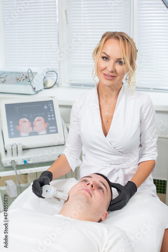 Photo of a young beautiful cosmetologist conducting a laser facial hair removal procedure with a modern device to a man lying on a couch in a cosmetology office