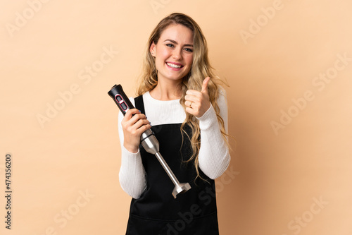 Young brazilian woman using hand blender isolated on beige background giving a thumbs up gesture