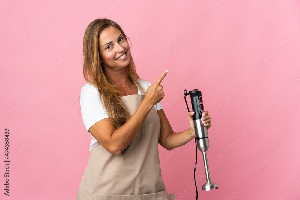 Middle age woman using hand blender isolated on pink background pointing back Middle age woman using hand blender isolated on pink background pointing back