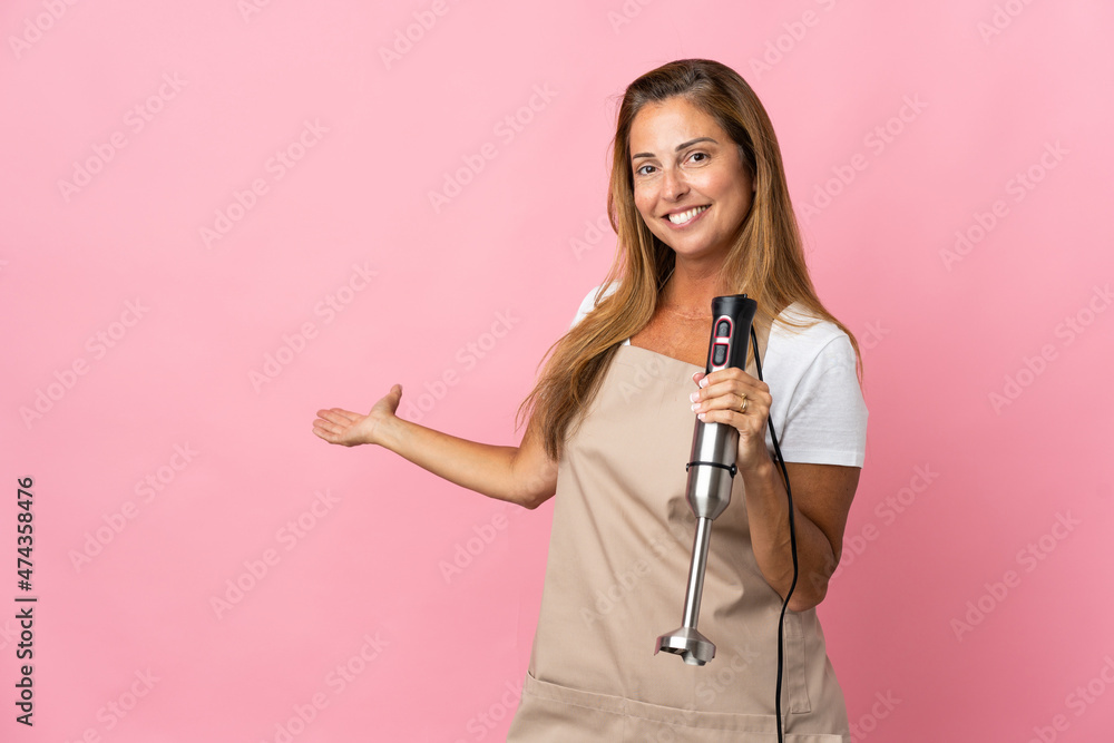 Middle age woman using hand blender isolated on pink background extending hands to the side for inviting to come Middle age woman using hand blender isolated on pink background extending hands to the side for inviting to come