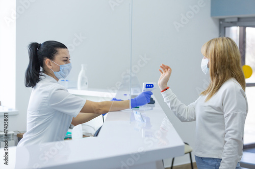 Shot of a female person being measured body temperature with a contactless thermometer by the employee of the reception desk of a modern hospital. Compliance with measures for the non proliferation of photo