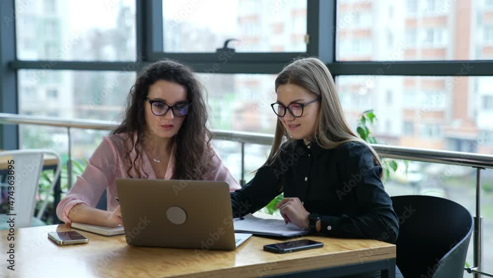 Two women colleagues discuss something in computer in office. Female teammates wearing glasses work in front of laptop. Two women colleagues discuss something in computer in office. Female teammates wearing glasses work in front of laptop.