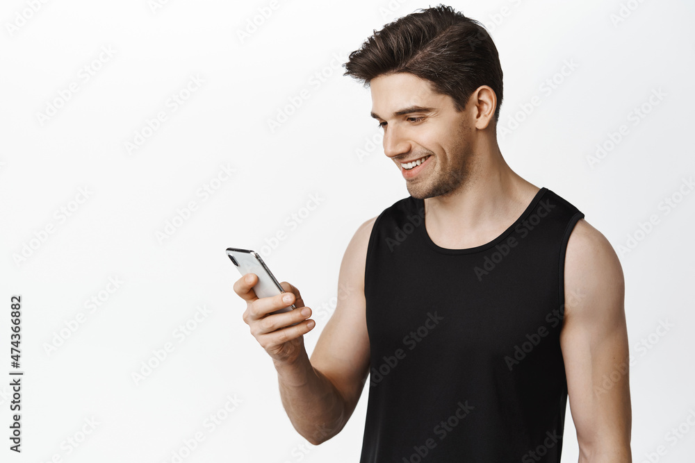 Sport and people concept. Young male athlete with fit and healthy body, looking at mobile phone, using application on smartphone, standing over white background Sport and people concept. Young male athlete with fit and healthy body, looking at mobile phone, using application on smartphone, standing over white background