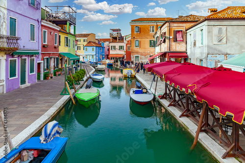 Burano Island, Venice, Italy. View of a canal in the town of Burano with the brightly colored houses famous all over the world. There are colored boats in the canal. Sunny blue sky with clouds. © Giulio Benzin