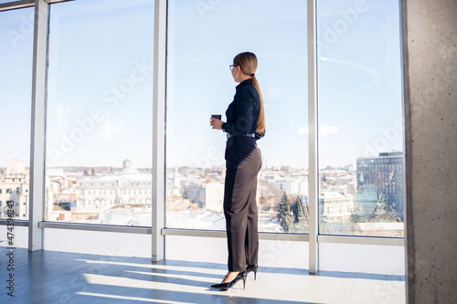 Confident business woman boss standing in modern office or hotel window enjoying big city view, woman leader, business owner thinking about future success, planning new opportunities,