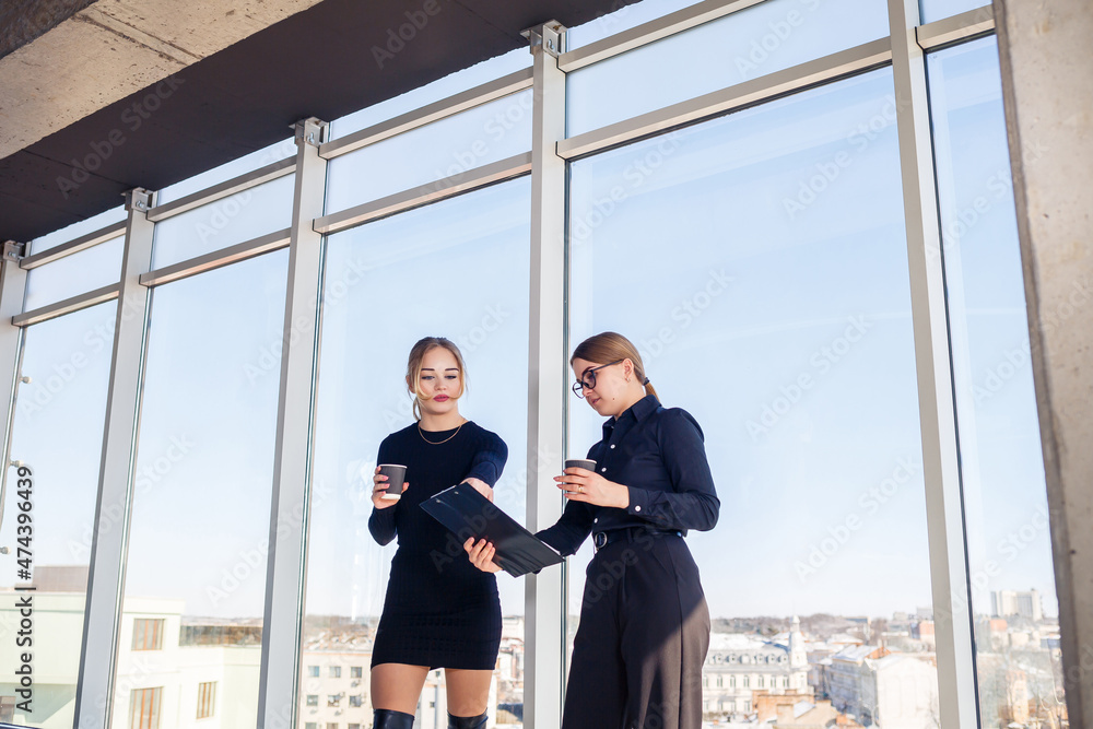 Two successful female managers talking about a joint project while standing by the window in the office. Young women economists, dressed in formal attire, talk during their breaks and drink coffee. Two successful female managers talking about a joint project while standing by the window in the office. Young women economists, dressed in formal attire, talk during their breaks and drink coffee.