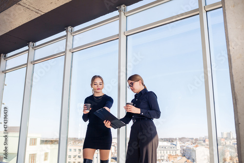 Two successful female managers talking about a joint project while standing by the window in the office. Young women economists, dressed in formal attire, talk during their breaks and drink coffee.
