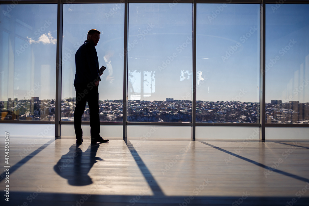 Rear view of a businessman looking out of a large window overlooking the city. He has a phone in his hands. Horizontal view. Selective focus