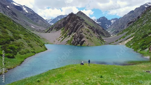 A drone flies over a turquoise mountain lake, two travelers next to a pond. the lake is surrounded by mountains and hills of konyon with small and large stones and various green vegetation.