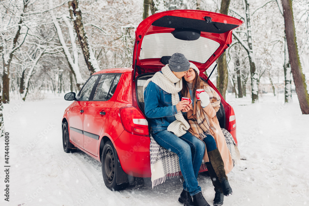 Loving couple sitting in car trunk drinking hot tea in snowy winter forest. People relaxing outdoors during road trip