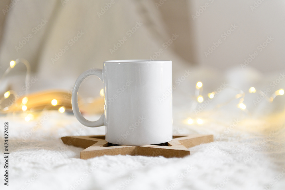 White mug mockup with blank copy space. Coffee cup on a beige knited background with opened book and christmas garland lights, cozy winter day at hugge home White mug mockup with blank copy space. Coffee cup on a beige knited background with opened book and christmas garland lights, cozy winter day at hugge home