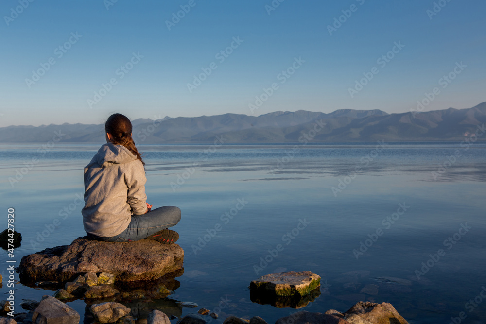 Quiet sunset over the river. The girl is sitting on a big stone, summer calm evening.