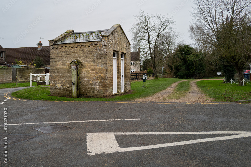 Roadside view of the oldest public toilet building in the UK, seen next to a village road junction. Still in use and showing a derelict water pump on the wall.