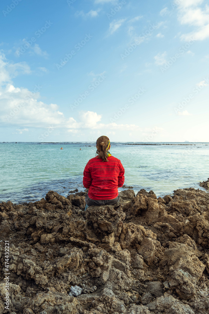 Mujer joven de pelo castaño vestida con una chaqueta roja sentada en unas rocas en una isla mirando al horizonte