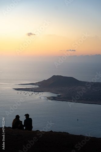 Silueta de dos jóvenes sentados mirándose con luz de atardecer y una isla en el océano al fondo © Anta