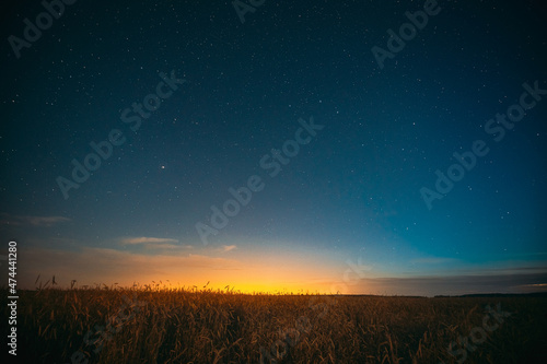 Night Starry Sky With Glowing Stars Above Countryside Landscape. Light Cloudiness Overcast Above Rural Field Meadow In Summer