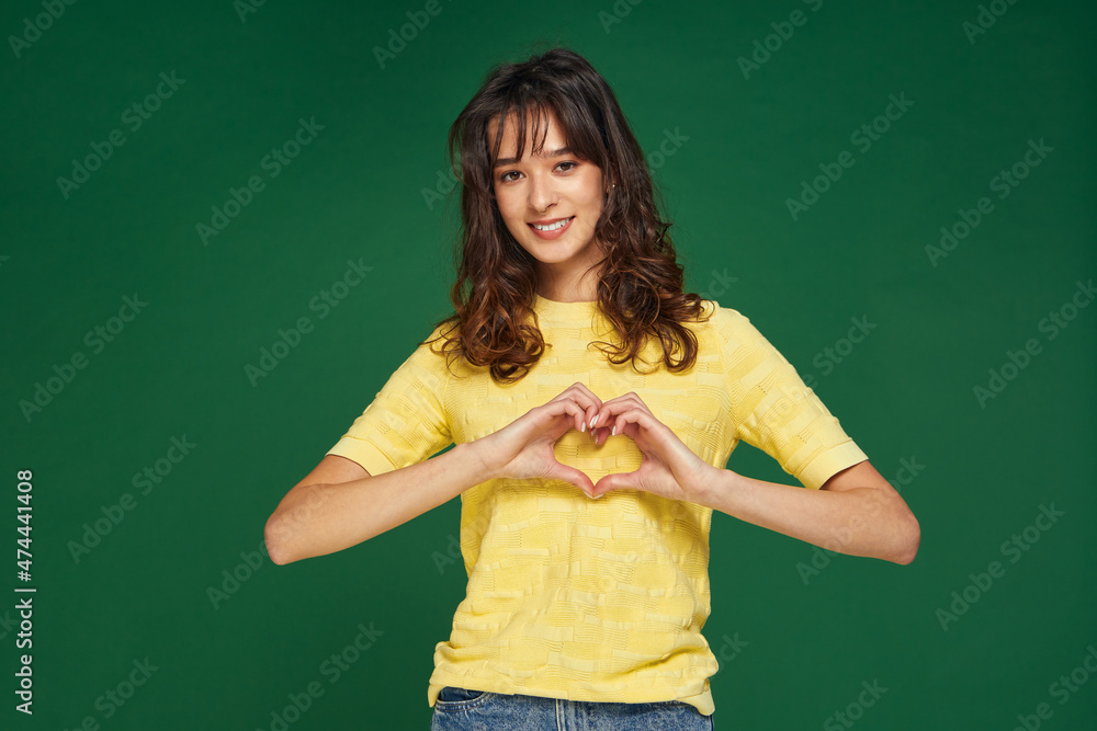 Smiling romantic young girl showing heart gesture, love valentine sign, feeling sympathy on green studio background Smiling romantic young girl showing heart gesture, love valentine sign, feeling sympathy on green studio background