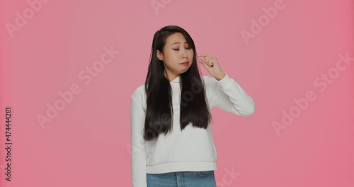 Upset young asian girl showing with fingers small size gesture, little measurement on pink studio background