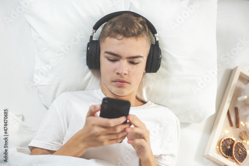 A nice teenage boy in white pajamas and headphones lies in bed with a smartphone in his hands. Dependence on gadgets. Lazy morning of vacation. A boy listens to music in bed.