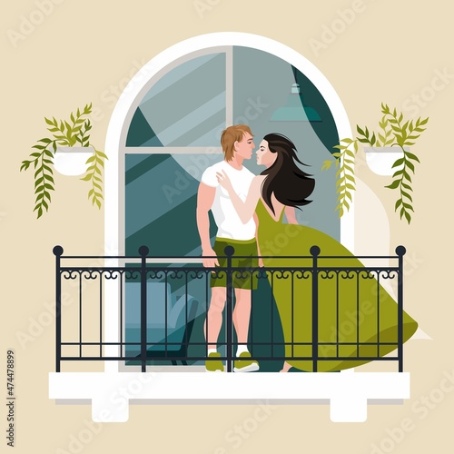 A couple in love stands and kisses on the balcony against the backdrop of a ponderous window with an arch. A balcony with green potted plants adorns the architecture of the building.