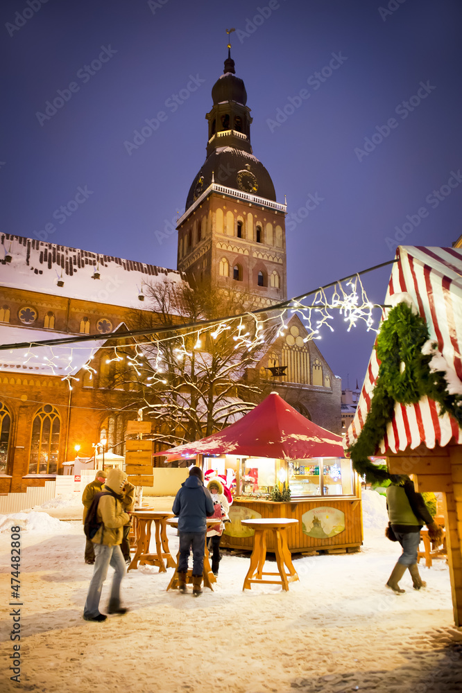 Fototapeta premium Winter evening on the main square of the city, street by evening lights, before Christmas, New Year. Christmas Fair. Catholic cathedral, church, tower. Riga, Latvia.