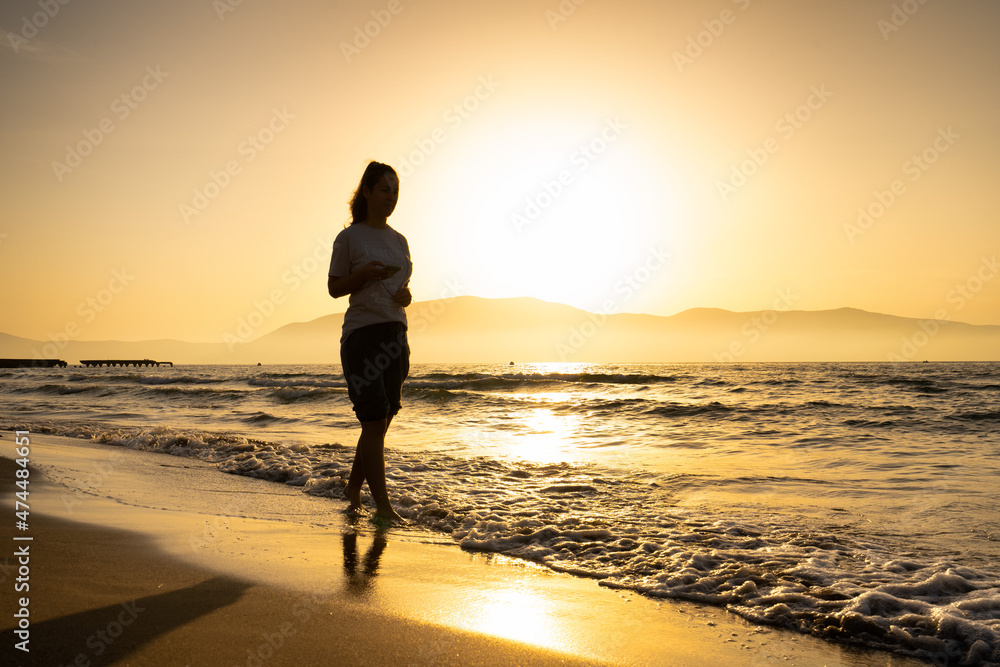 Pretty woman silhouette walks at seaside surf in golden sunset light. Girl having fun on ocean beach on summer