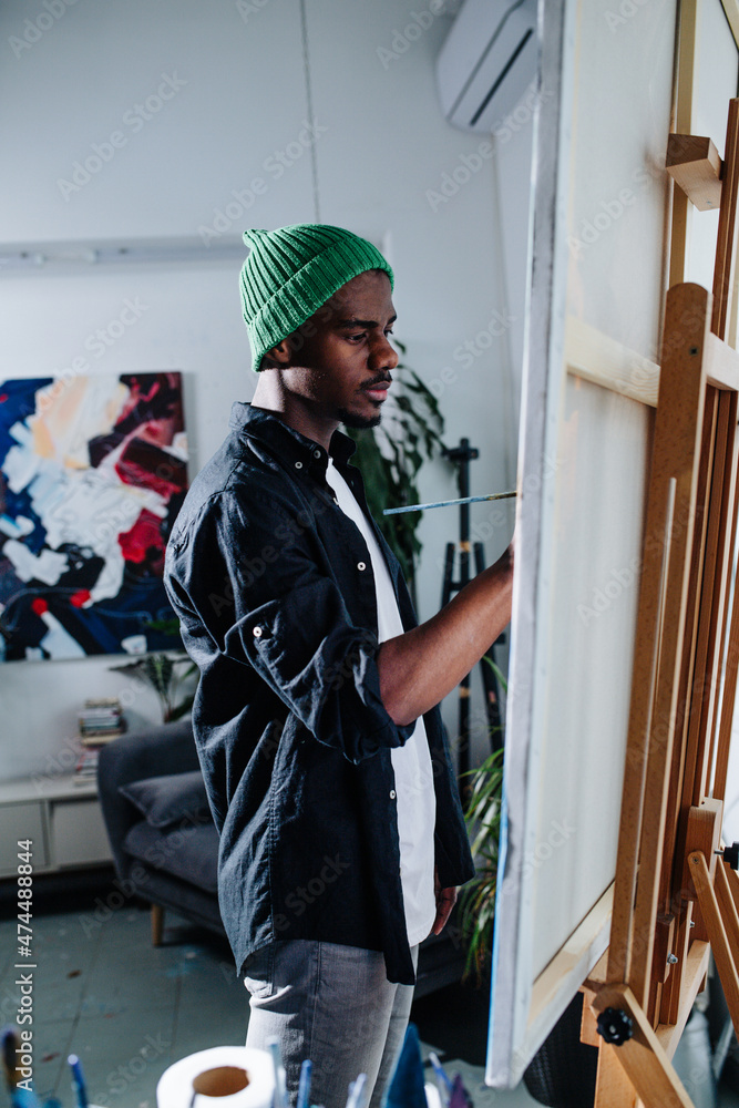 Focused peaceful black man painting on an easel inside of his apartment