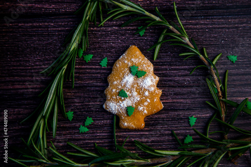 Cookies in the form of a Christmas tree, sprinkled with powdered sugar and small Christmas trees on top, lies on a dark wooden background surrounded by grained rosemary. High quality photo photo