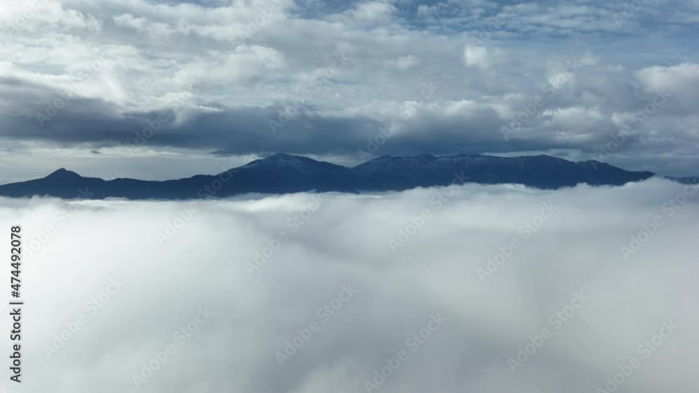 Flying above the clouds, stunning aerial view of a mountain range in the distance surrounded by clouds during a winter day in Italy. Flying above the clouds, stunning aerial view of a mountain range in the distance surrounded by clouds during a winter day in Italy.