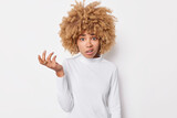 Photo of indignant curly haired woman raises hand shrugs shoulders feels skeptical being displeased wears casual turtleneck isolated over white background. Bothered questioned female model indoor