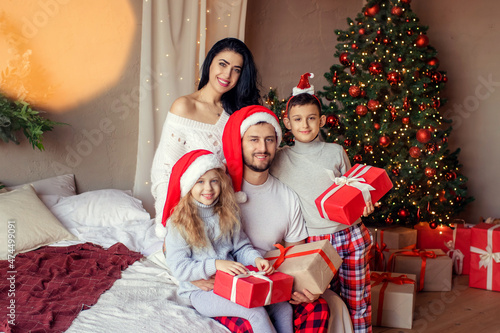 Portrait of young happy family parents and children with gift boxes on New Years Eve sitting on the bed near the decorated Christmas tree at home.