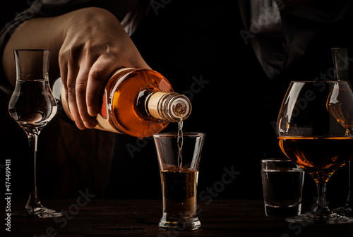The bartender pours golden tequila in shot glass on the old wooden bar counter. Vintage background in pub or bar, night mood. Place for text