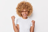 Happy triumphing young woman with curly hair clenches fists celebrates success glad to achieve goals dressed in casual t shirt isolated over white background. Ambitious successful female model