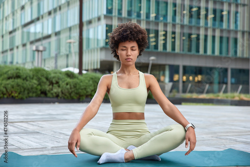 Full length shot of peaceful curly haired female model with curly hair meditates and practices yoga sits crossed legs on mat controlls emotions poses against urban background. Strees free concept