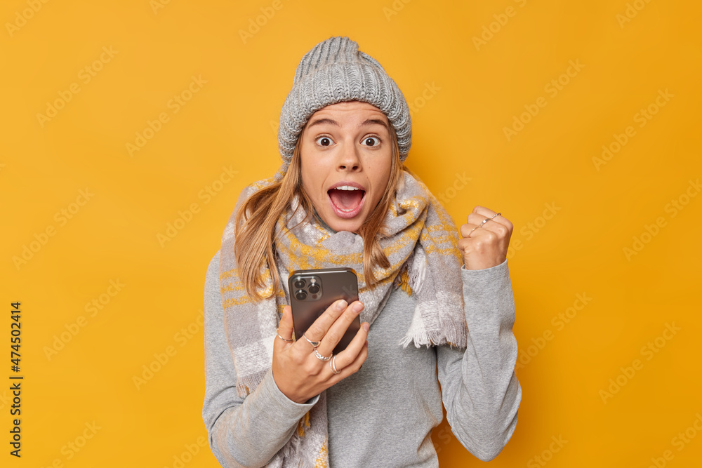 Sweet taste of success. Positive excited woman gained her daily app in goal holds mobile phone clenches fist exclaims loudly wears knitted hat jumper and scarf around neck isolated over yellow wall