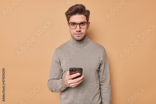 Serious young man distracted from screen after watching video holds mobile phone looks confidently at camera uses modern gadget wears eyeglasses and turtleneck isolated over beige background