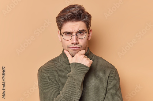 Photos Portrait of serious skeptic man holds chin looks attentively at camera feels self confident considers something wears round spectacles and casual sweater isolated over brown background
