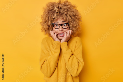 Scared frightened curly haired woman bites lips feels panic being worried about something trembles from fear notices scary thing wears transparent eyeglasses and jumper isolated over yellow background © Wayhome Studio