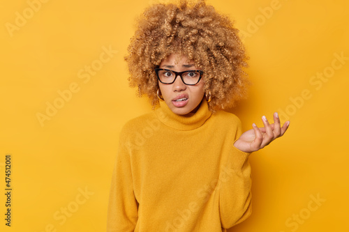 Indoor shot of indignant curly haired woman shrugs shoulders raises palm with displeased expression purses lips wears spectacles and jumper isolated over vivid yellow background feels indecisive. photo