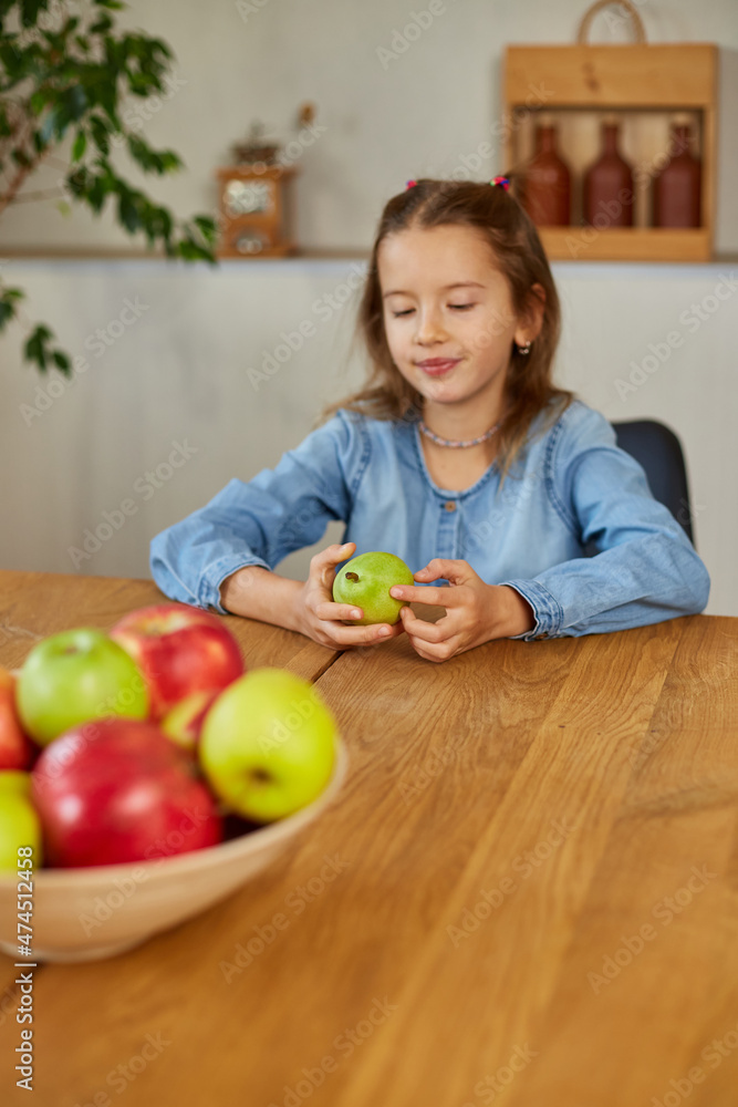 Fototapeta premium Happy little girl takes a pear from the bowl with variety fruits on table at home