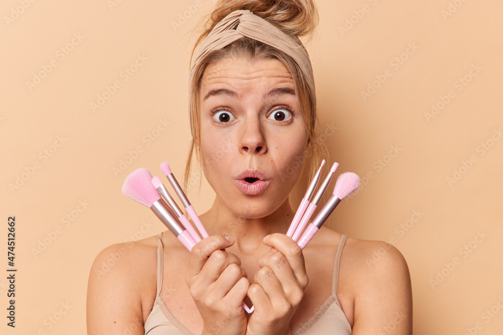Shocked young European woman holds cosmetic brushes undergoes beauty routine uses new tools reacts on something astonishing wears headband and casual t shirt isolated over brown studio background Shocked young European woman holds cosmetic brushes undergoes beauty routine uses new tools reacts on something astonishing wears headband and casual t shirt isolated over brown studio background