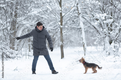 A young man in a gray down jacket plays with a Yorkshire terrier dog in a winter forest