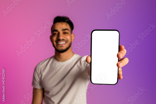 Smiling young Arab man showing smartphone with blank screen, recommending website or ad in neon light, mockup