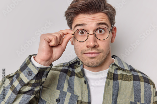 Photo of surprised man stares through spectacles wears checkered shirt cannot believe his eyes has stubble reacts on astonishing news isolated over white background. Human reactions concept.